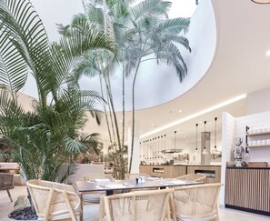 Un espace repas moderne, doté d’un haut plafond et baigné de lumière naturelle, avec une table centrale entourée de chaises, de grandes plantes en pot et un coin cuisine en arrière-plan.