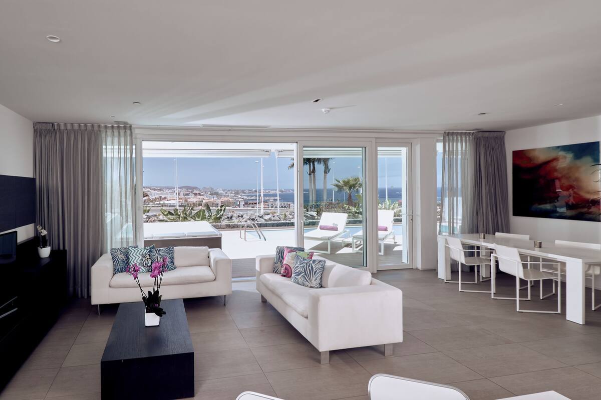 A modern living room with a large window, white sofas, a dining area, and a view of the city and sea.