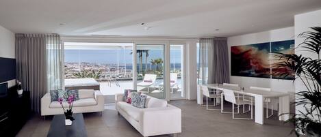 Un salon moderne doté d’une grande fenêtre, de canapés blancs, d’un coin repas et offrant une vue sur la ville et la mer.