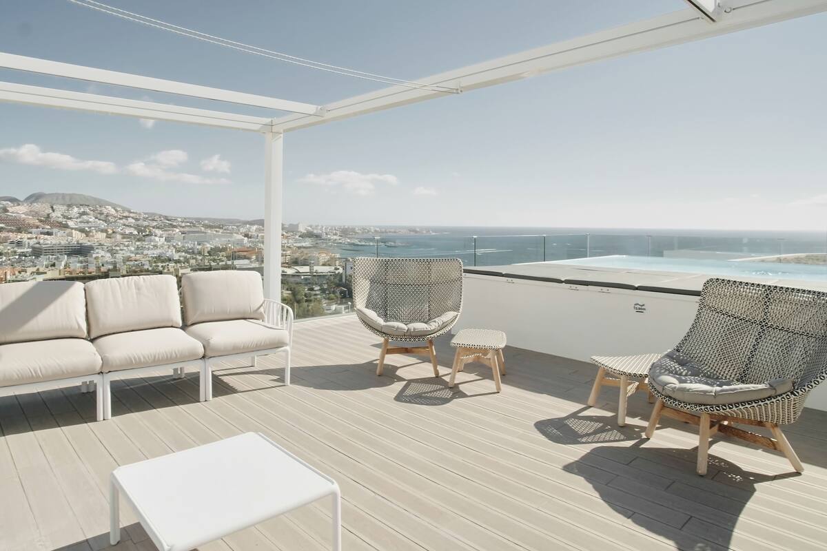 A rooftop terrace with a white sofa, two wicker chairs, and a small table. A pool is visible in the background.