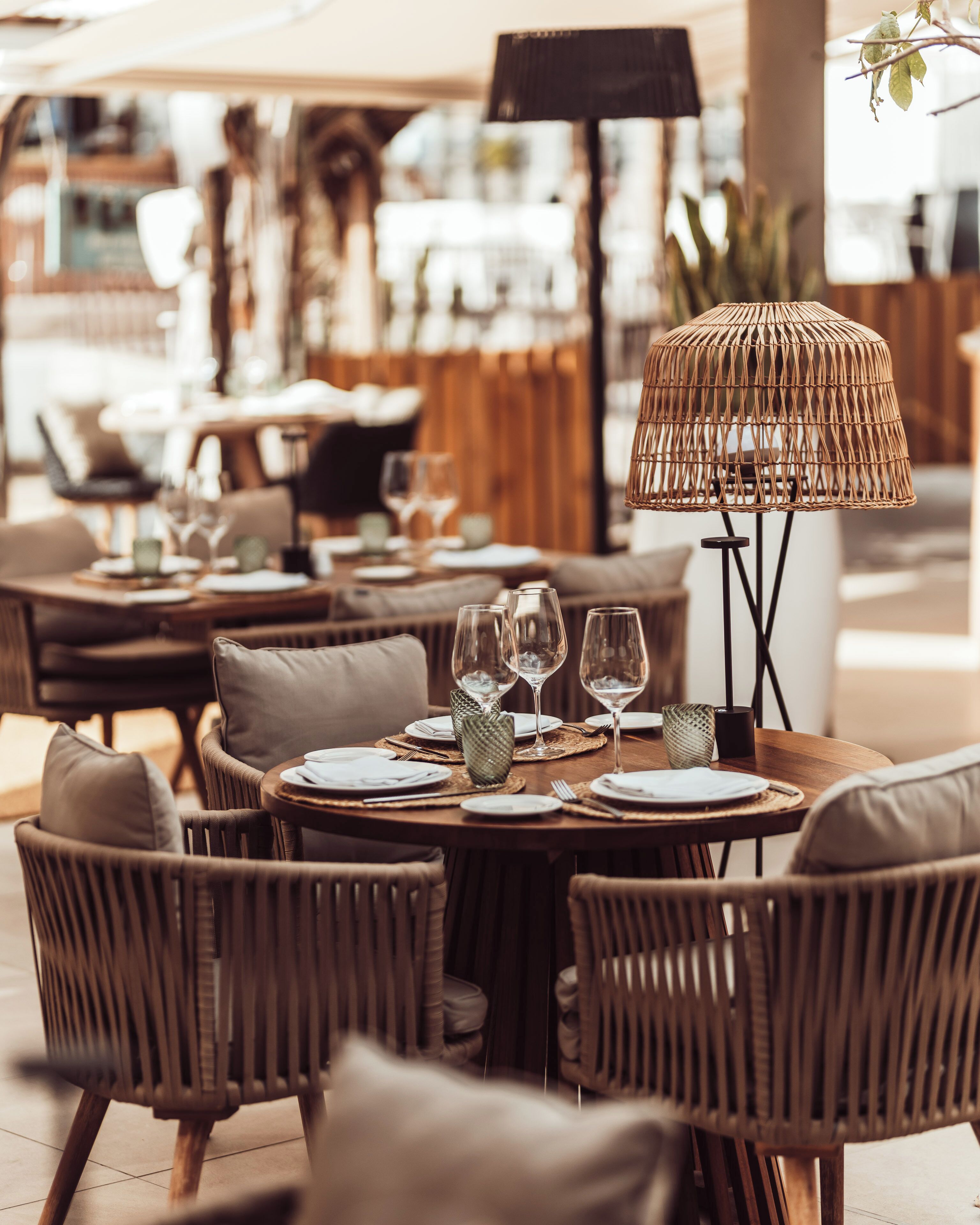 An outdoor dining area with wicker furniture and a woven lamp. Tables are set with plates, glasses, and cutlery.
