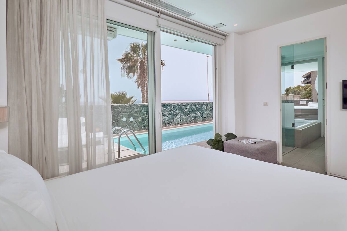 A hotel room with a large bed, a TV, a small table, and a view of a pool and ocean.