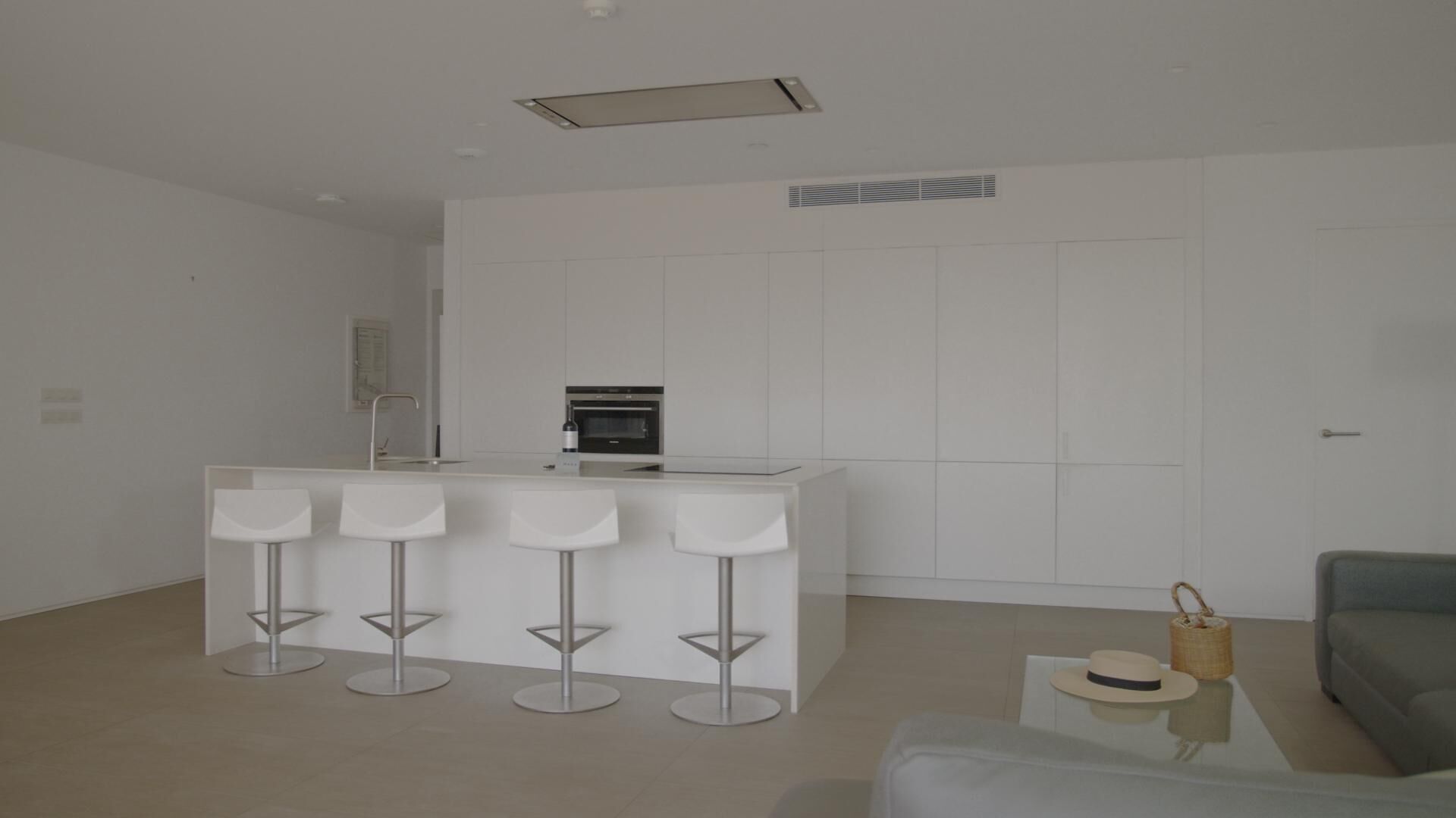 A modern kitchen with a central island, four bar stools, a grey sofa, and a small side table with a hat and basket.