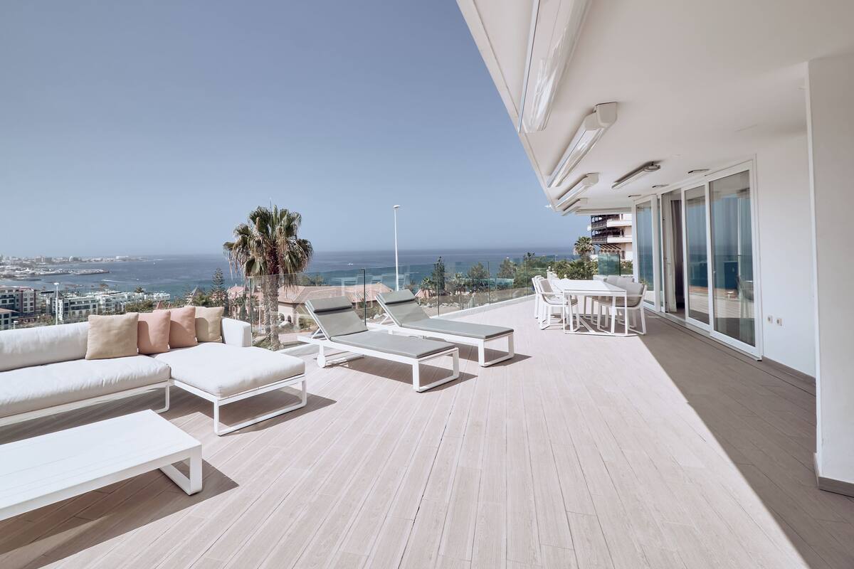 A sunlit outdoor terrace with a sofa, lounge chairs, and a table, overlooking a coastal cityscape.