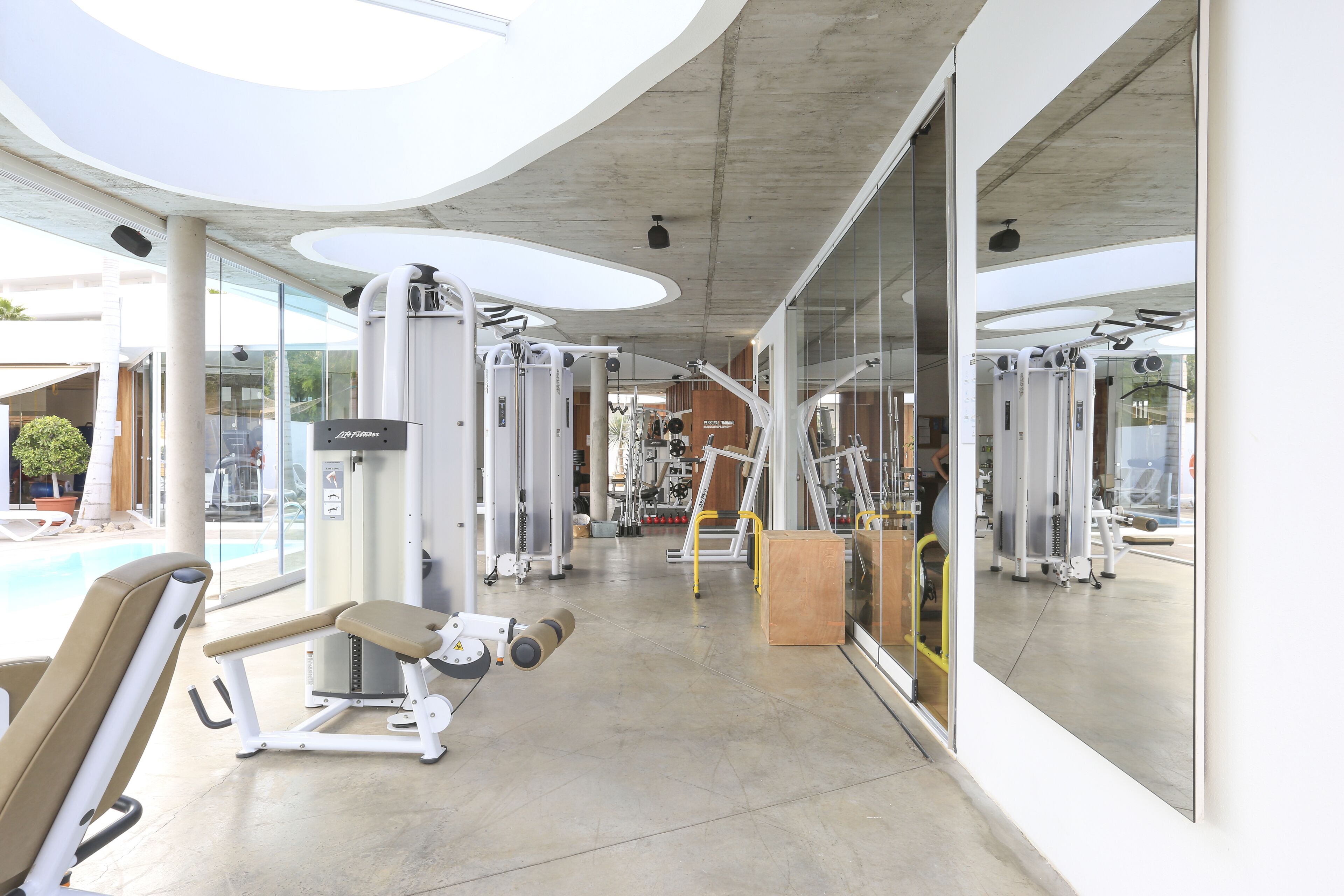 A modern gym with various exercise machines, large windows, and a pool area visible through the glass doors.