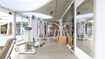 A modern gym with various exercise machines, large windows, and a pool area visible through the glass doors.