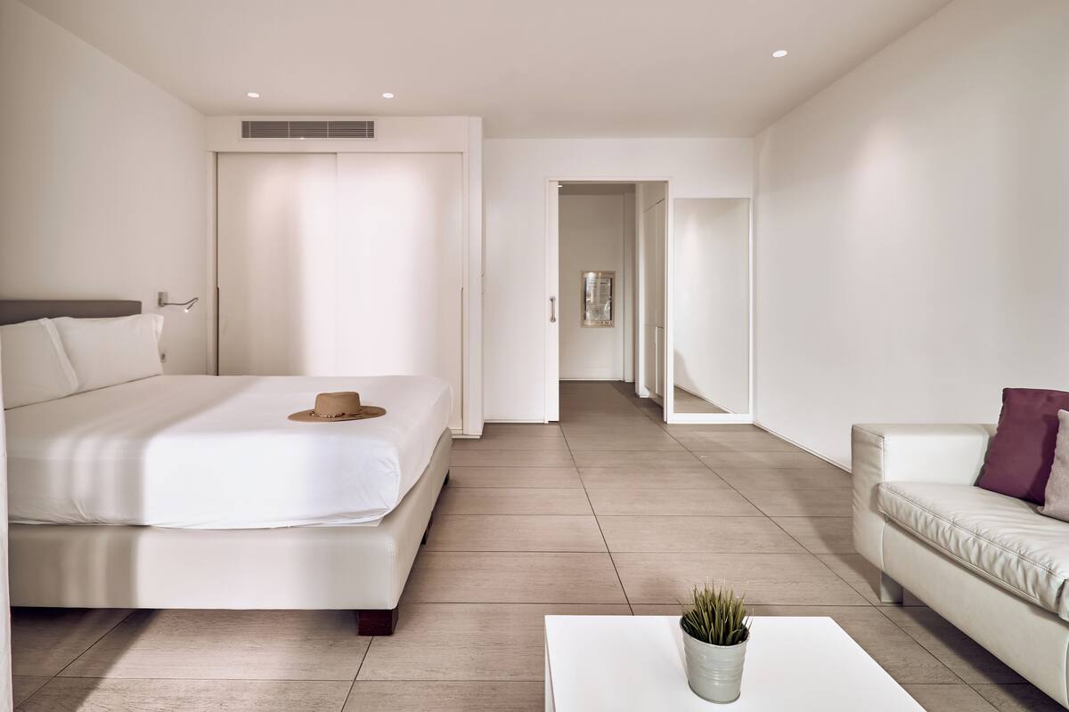 A modern hotel room with a bed, a sofa, a coffee table, and a hallway leading to another room.