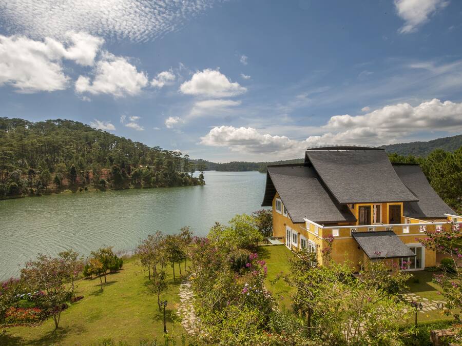 A house with a sloped roof overlooking a lake, surrounded by lush greenery and trees.
