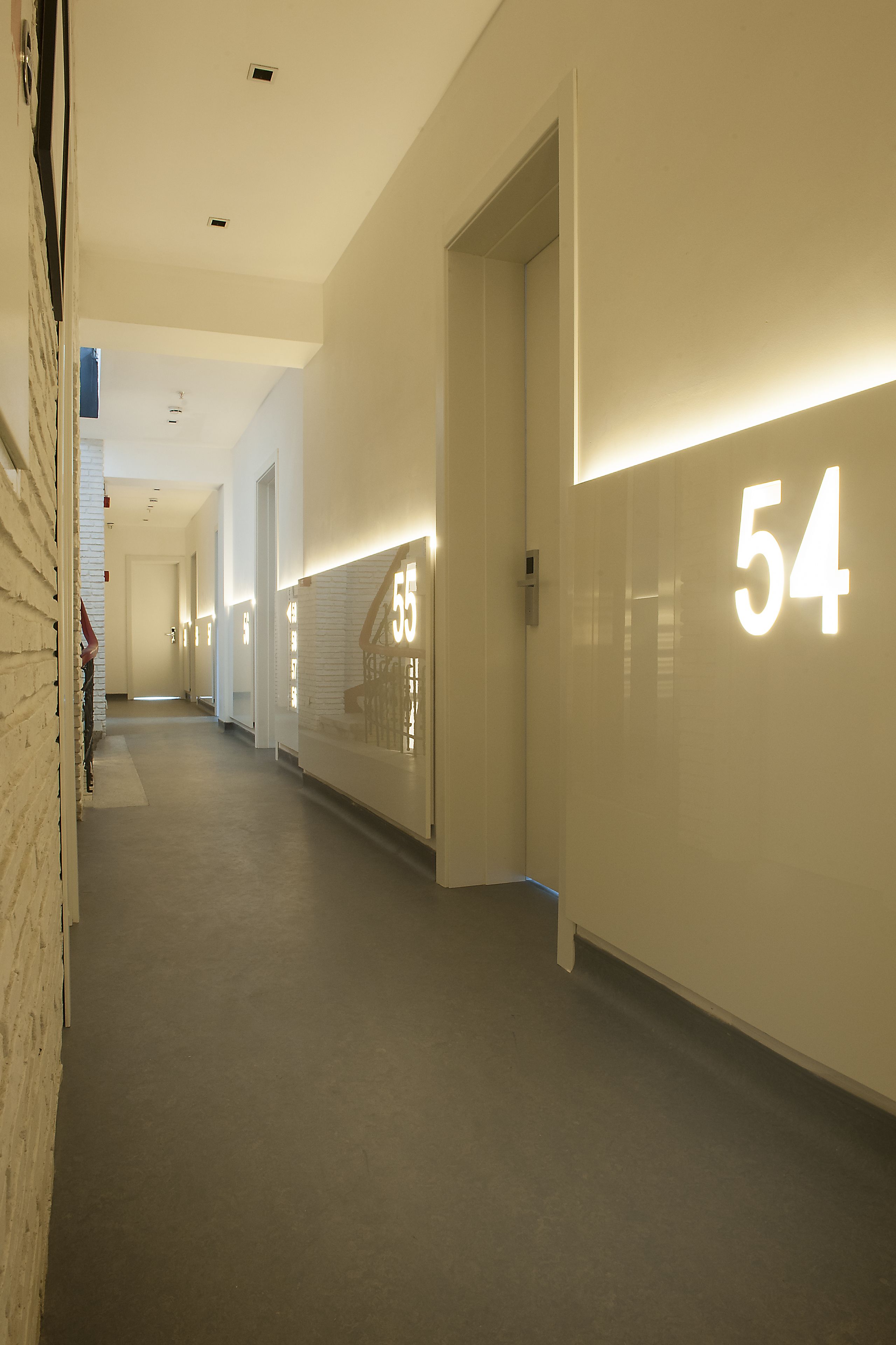 A corridor with illuminated room numbers, a clean floor, and a white brick wall on the left.
