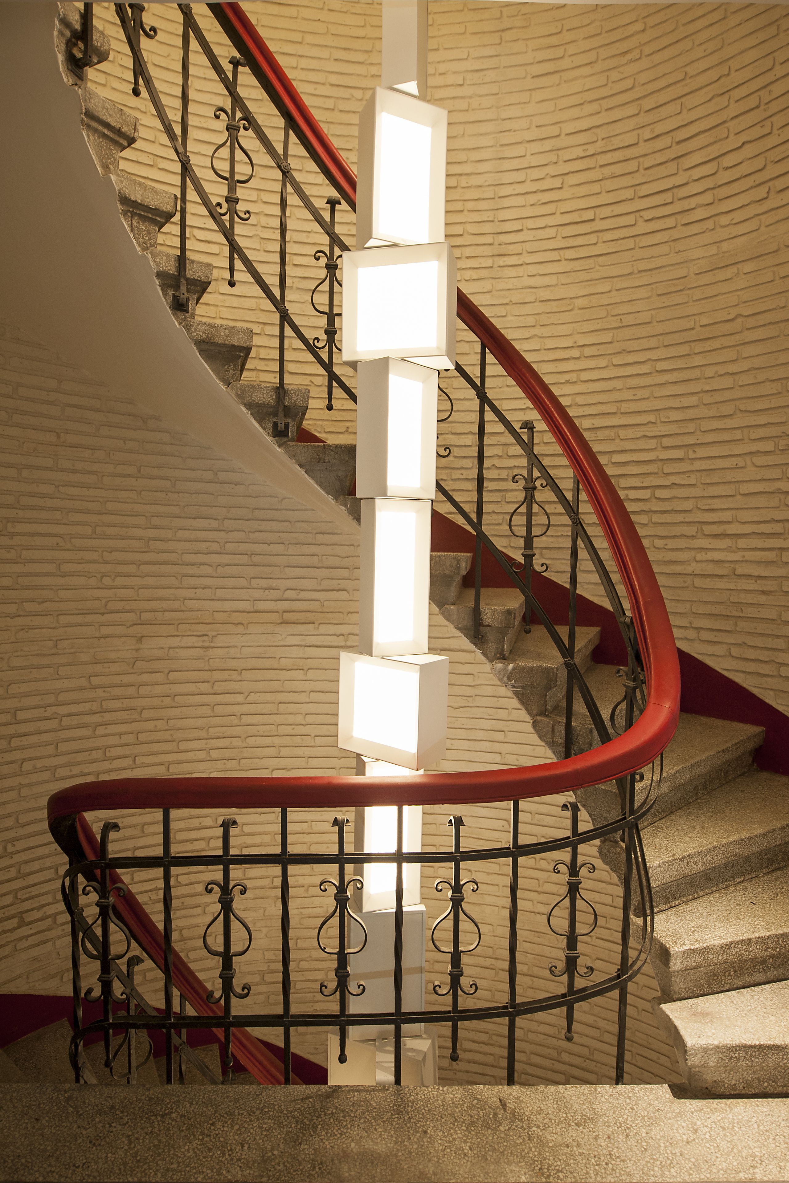 Eine Wendeltreppe mit roter Handlauf und schwarzen Spindeln, die zu einer Plattform mit weißer Wand und Ziegelmuster führt.