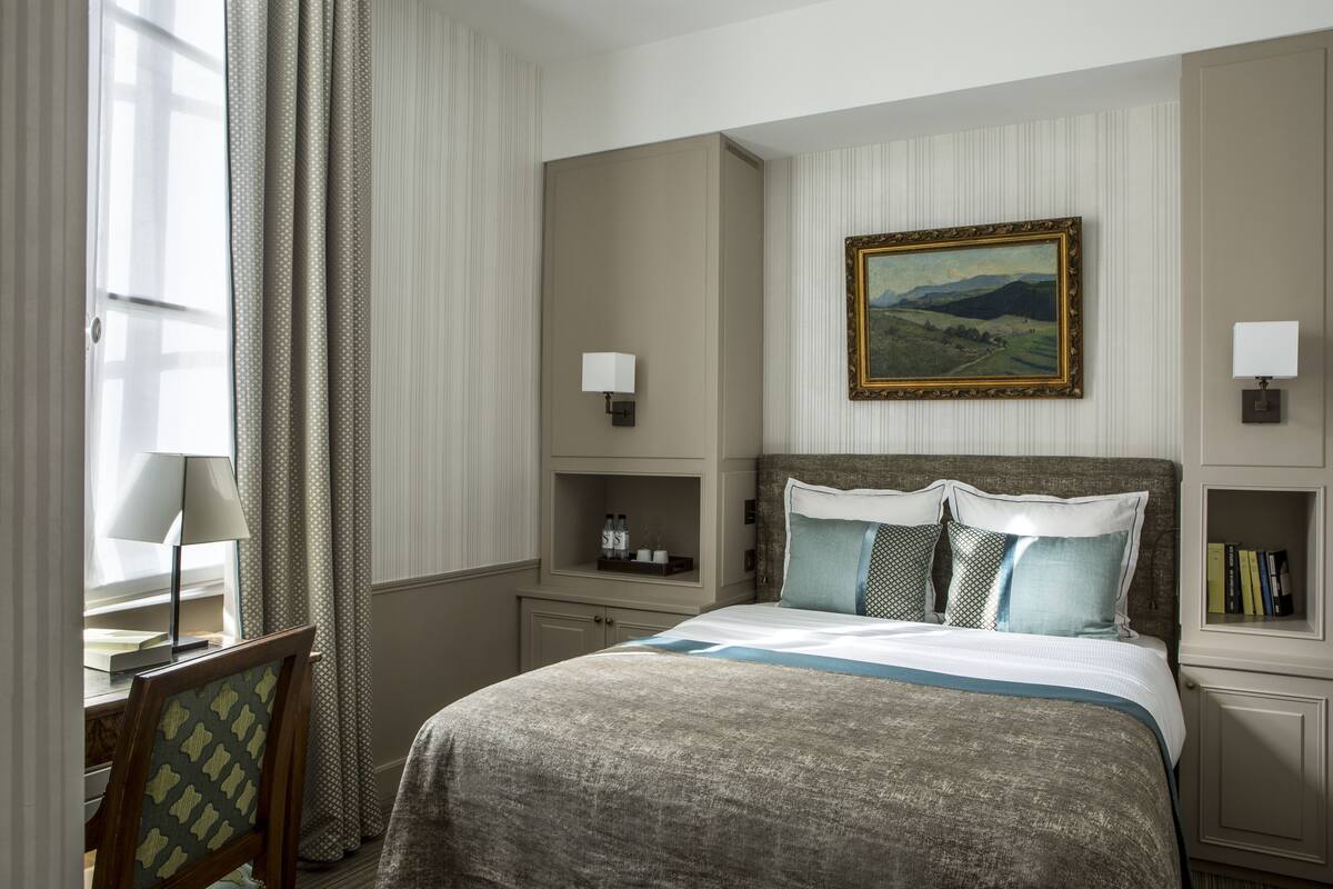 A hotel room with a large bed, two bedside tables with lamps, a framed painting on the wall, and a window with curtains.