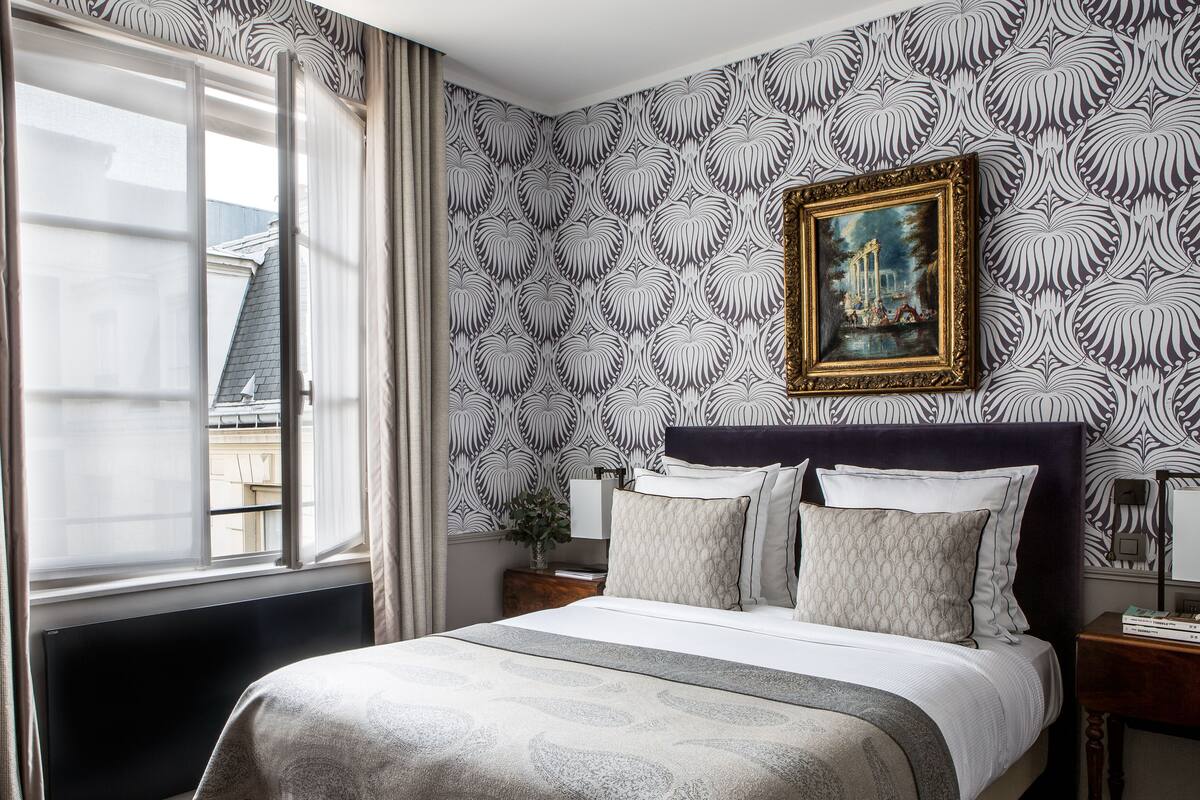 A bedroom with a large bed, a window with curtains, a patterned wallpaper, and a framed painting on the wall.