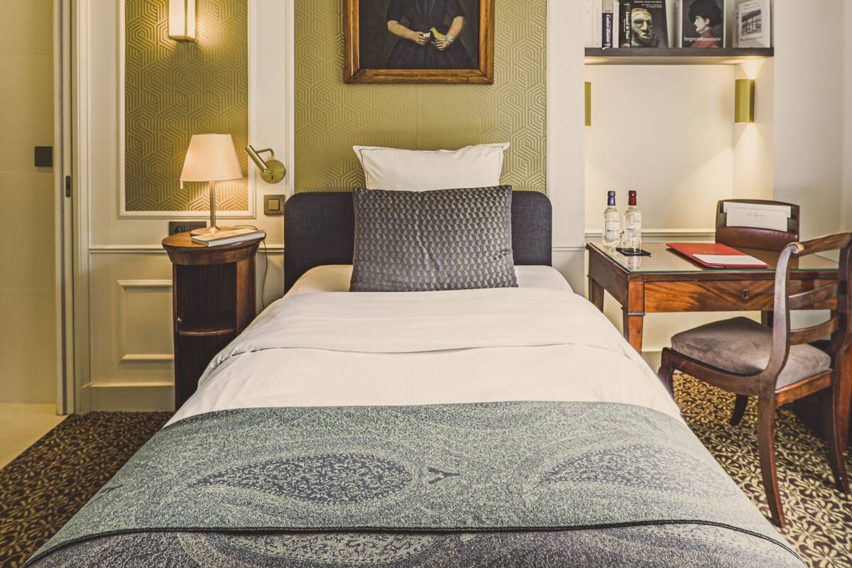 A hotel room with a bed, a desk, a chair, a painting on the wall, and a shelf with books and pictures.