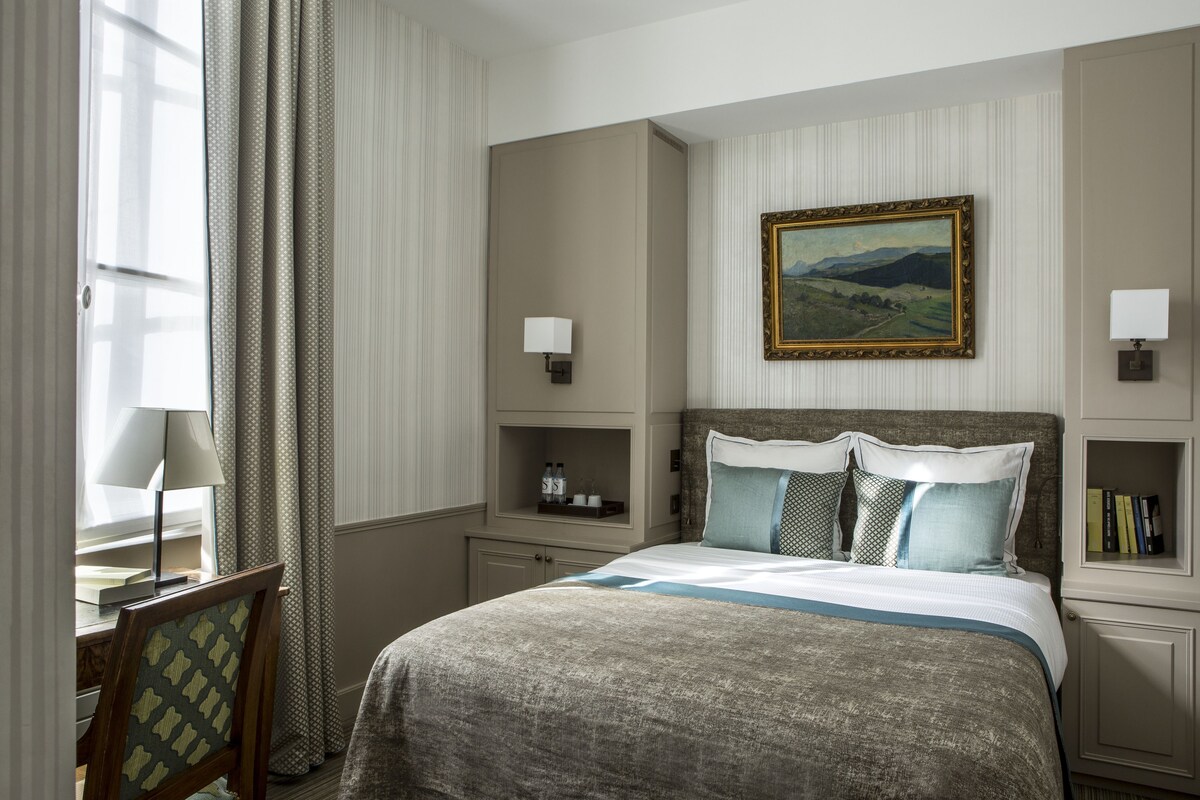 A hotel room with a bed, bedside tables, a nightstand, a framed painting, and a window with curtains.