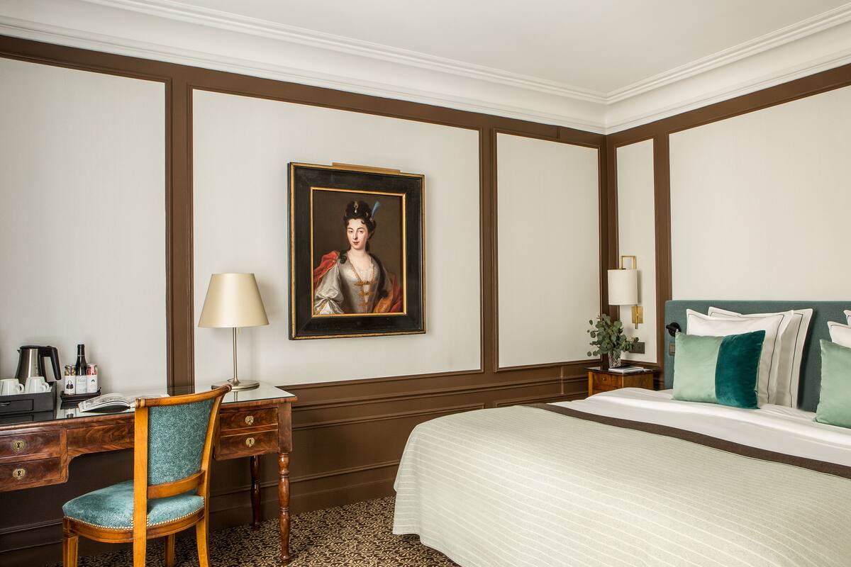 A hotel room with a bed, a desk, a chair, a lamp, and a painting on the wall.