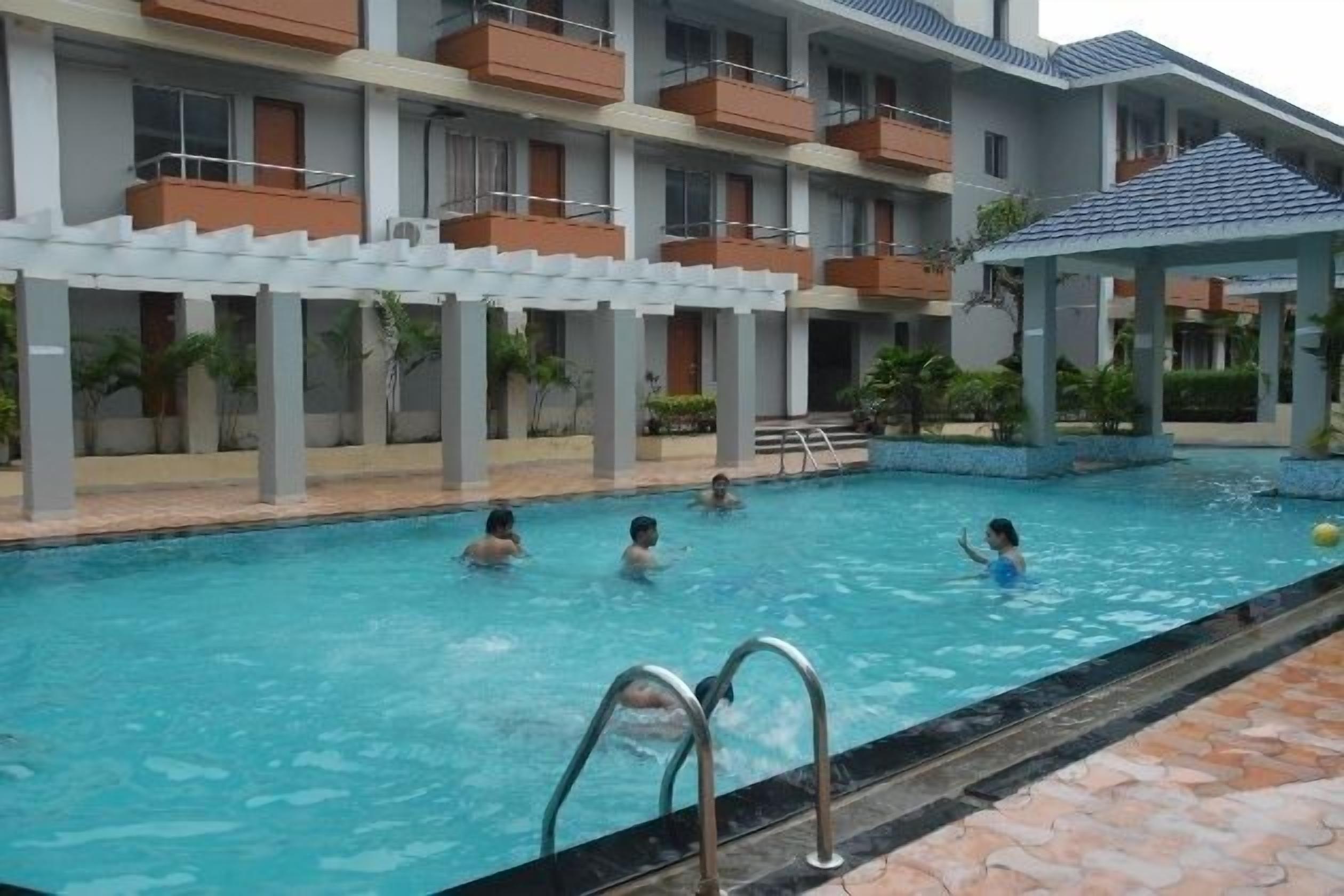 A swimming pool with people in the water, surrounded by a building with balconies and a covered area.
