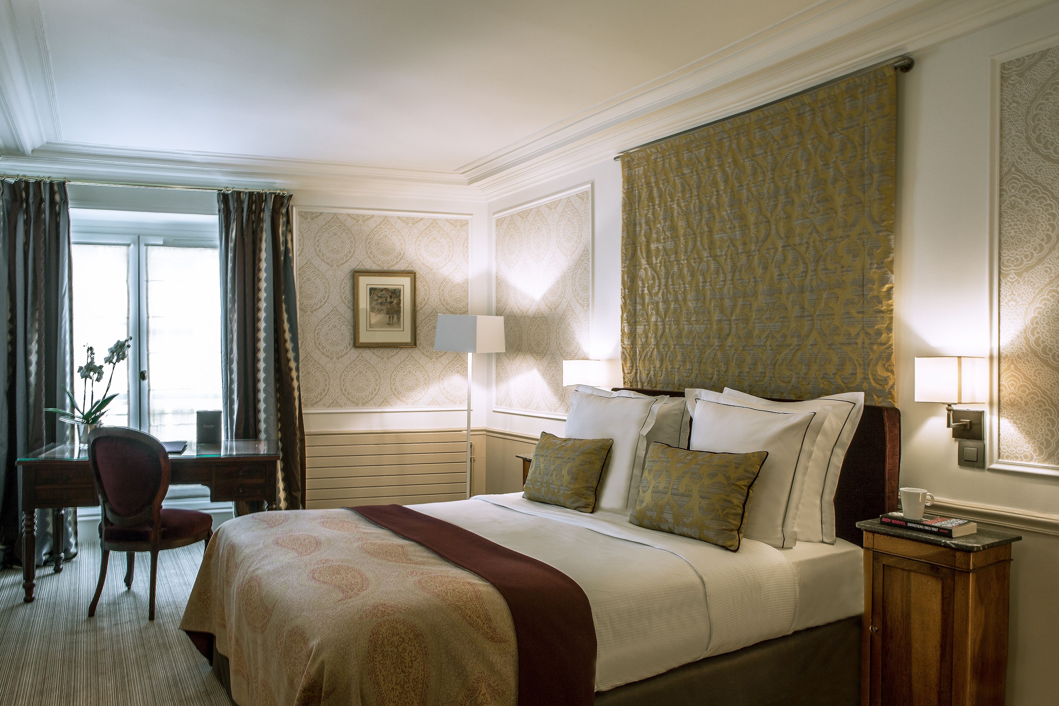A hotel room with a large bed, a desk, and a chair. There is a window with curtains, a lamp on the desk, and a picture on the wall.