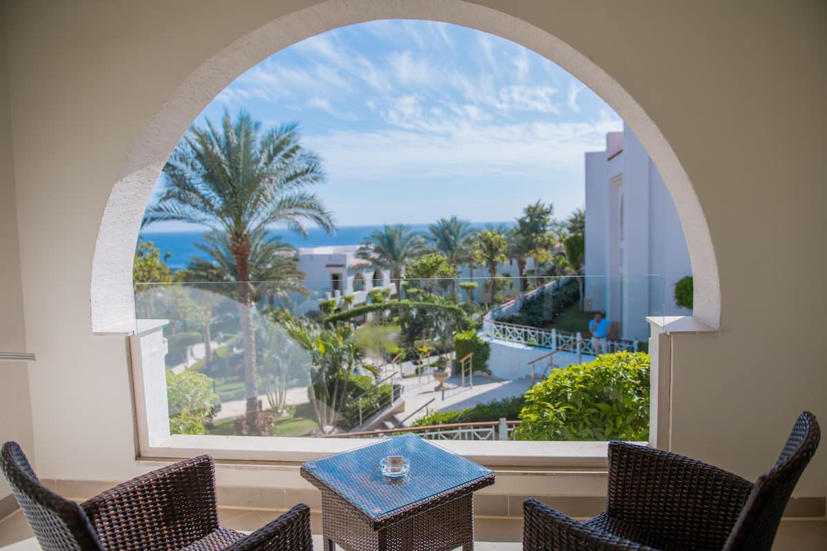 A balcony with wicker chairs and a small table, overlooking a landscaped area with palm trees and a view of the sea.