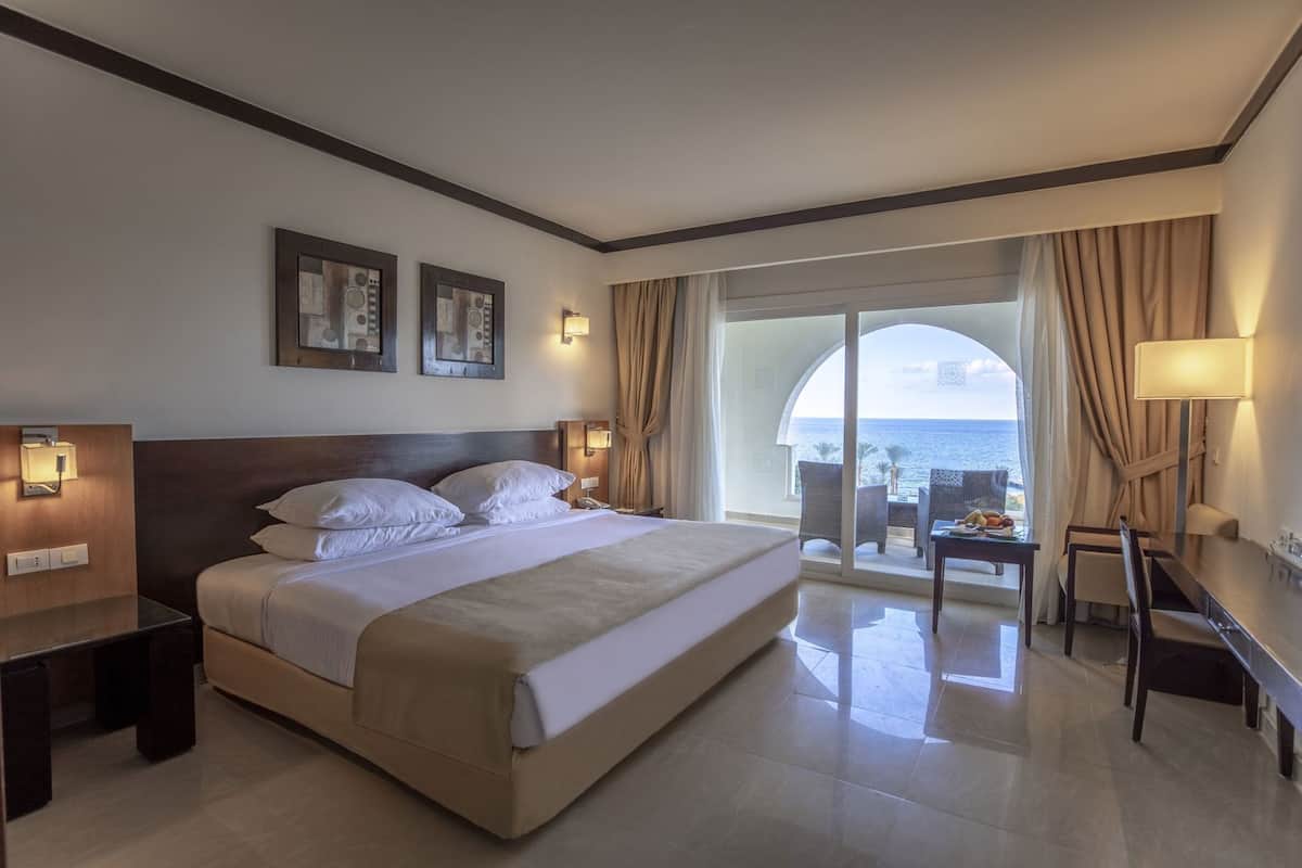 A hotel room with a large bed, a desk, a chair, and a balcony with a view of the sea.