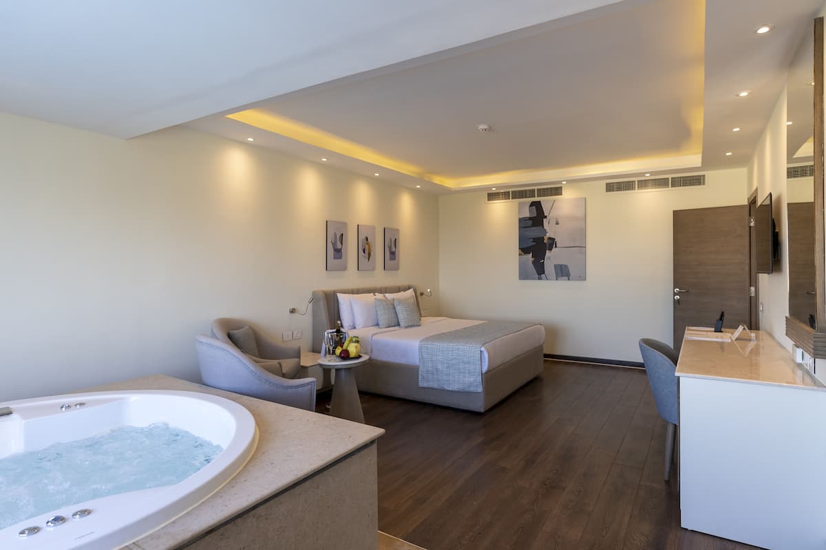 A modern hotel room with a large bed, a sitting area with a chair and a small table, a bathtub, and a desk with a mirror.