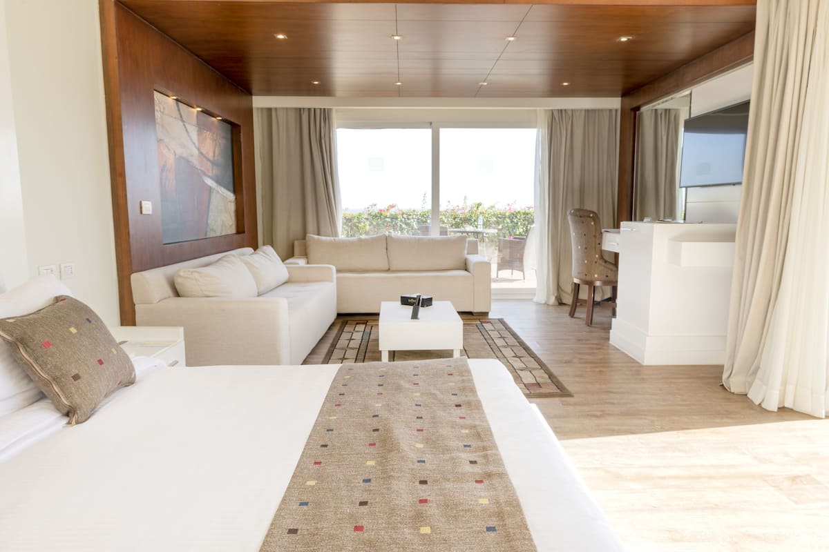 A modern hotel room with a large bed, a sofa, a small table, and a balcony with a view of greenery.