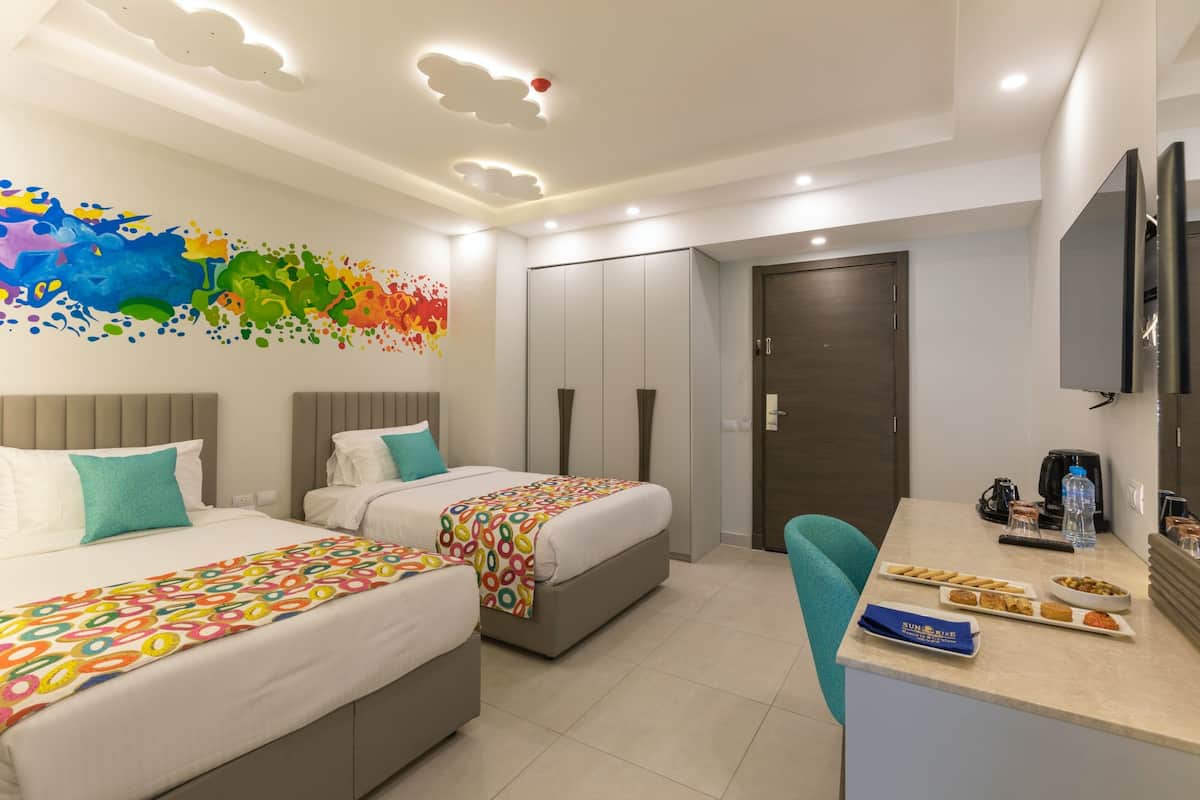 A modern hotel room with two beds, a desk with breakfast items, a TV, and a large abstract painting on the wall.