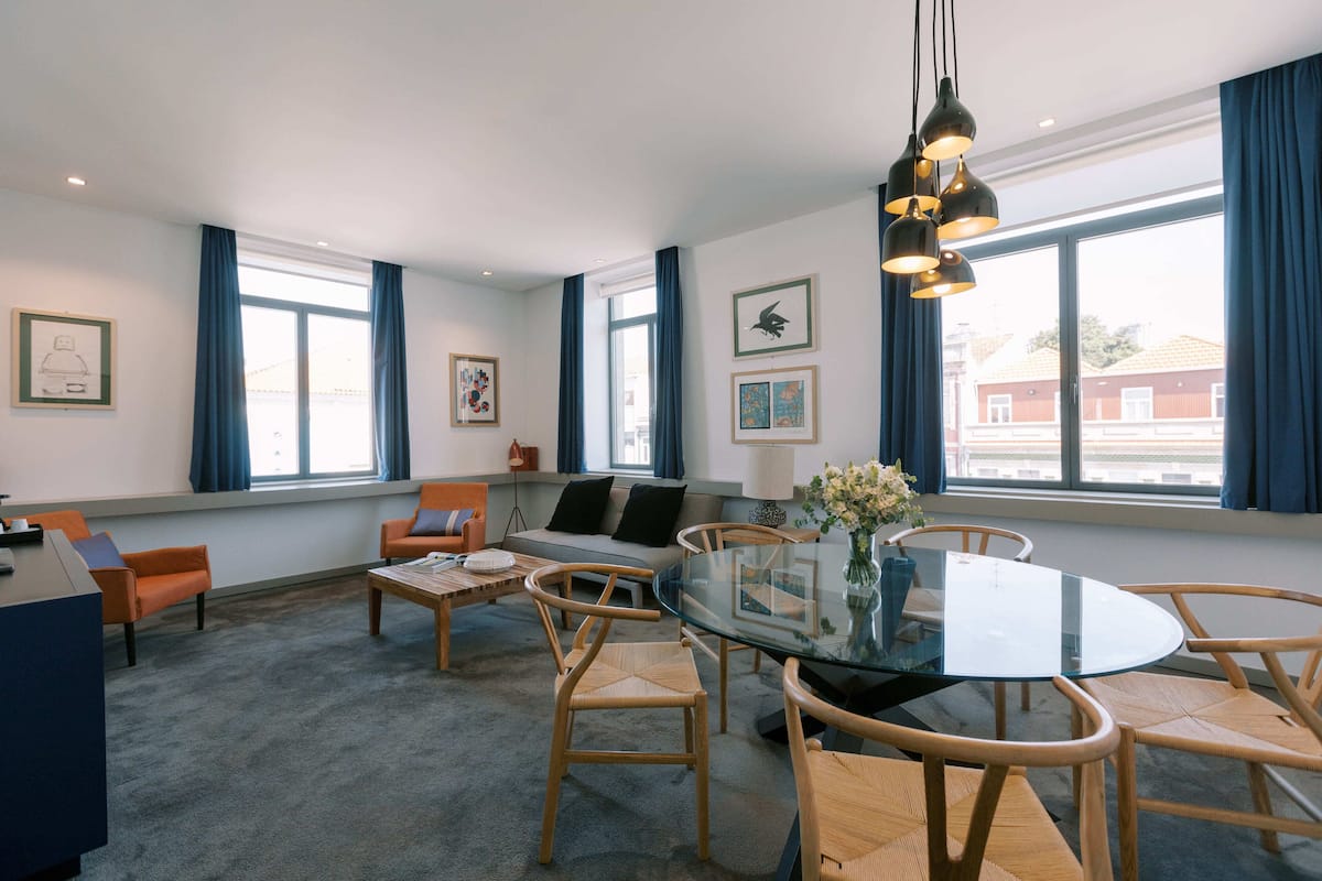 Un salon moderne avec une table à manger en verre, des chaises en bois, un canapé et une table basse. De grandes fenêtres sont ornées de rideaux bleus, des œuvres d’art décorent les murs et un luminaire suspendu illumine l’espace.