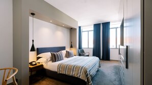 A modern hotel room with a large bed, bedside table with a lamp, a chair, and a TV mounted on the wall.