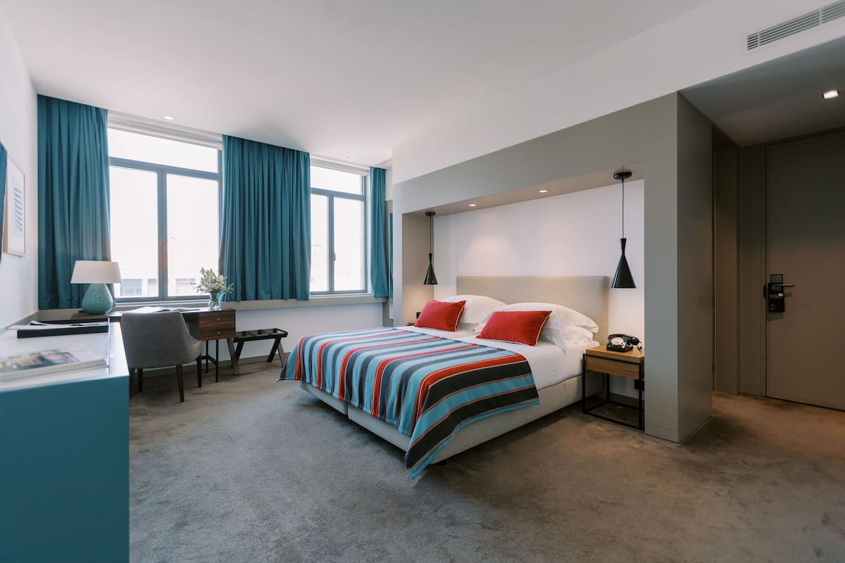 Une chambre d’hôtel moderne avec un grand lit, un bureau et une chaise. On y trouve une fenêtre avec des rideaux, une table de chevet avec une lampe et une porte.