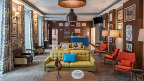 A hotel lobby with a yellow sofa, red chairs, a large black pendant light, and framed artwork on the walls.