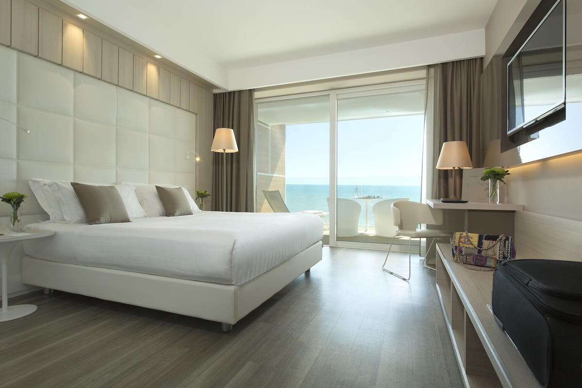 A modern hotel room with a large bed, a TV, a balcony with a view of the sea, and a small table with a lamp.
