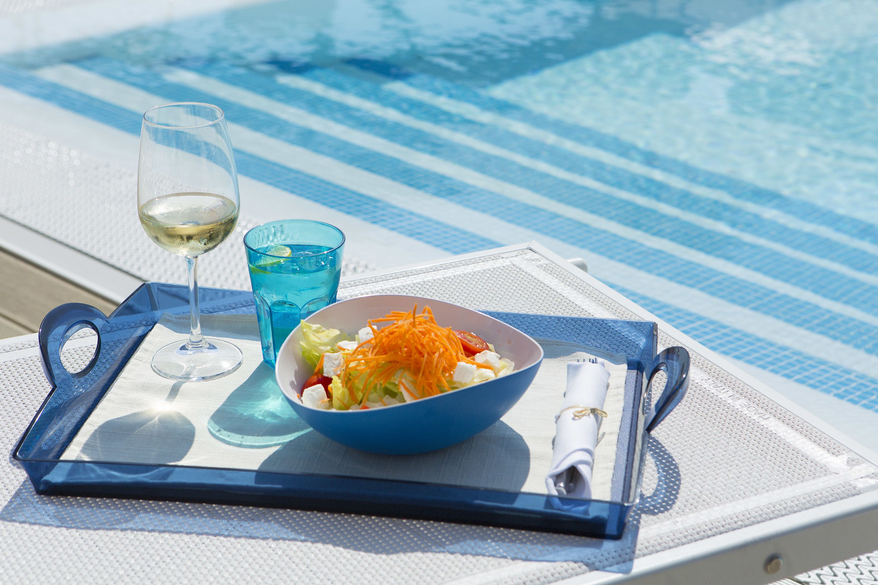 A dining table with a blue tray, a glass of white wine, a blue glass with a slice of lime, and a bowl of salad.