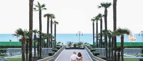 A couple walking on a bridge with palm trees and a view of the sea.