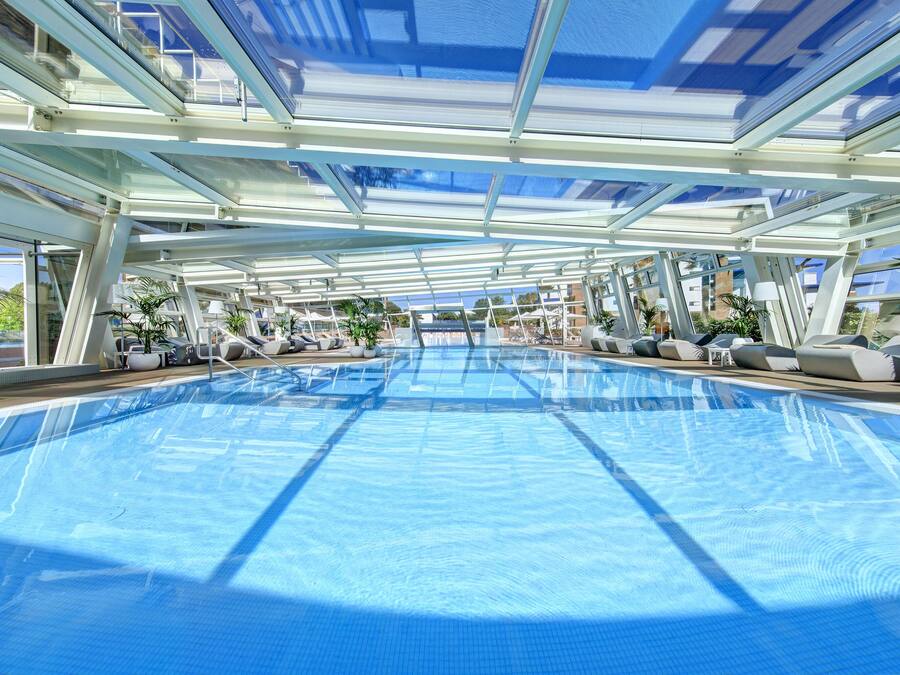 A large indoor swimming pool with clear blue water, surrounded by lounge chairs and potted plants.