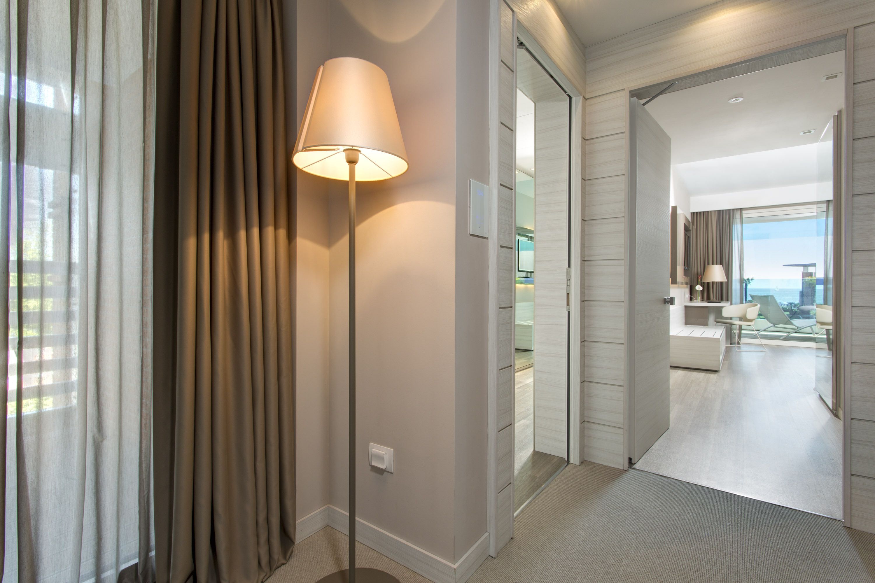 Un pasillo moderno de hotel con una lámpara, una puerta corredera que da a una habitación con vistas a la ciudad y una zona de estar con mesa y sillas.