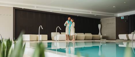 Un área interior de piscina moderna con una piscina cristalina, asientos de descanso y una pared oscura al fondo.