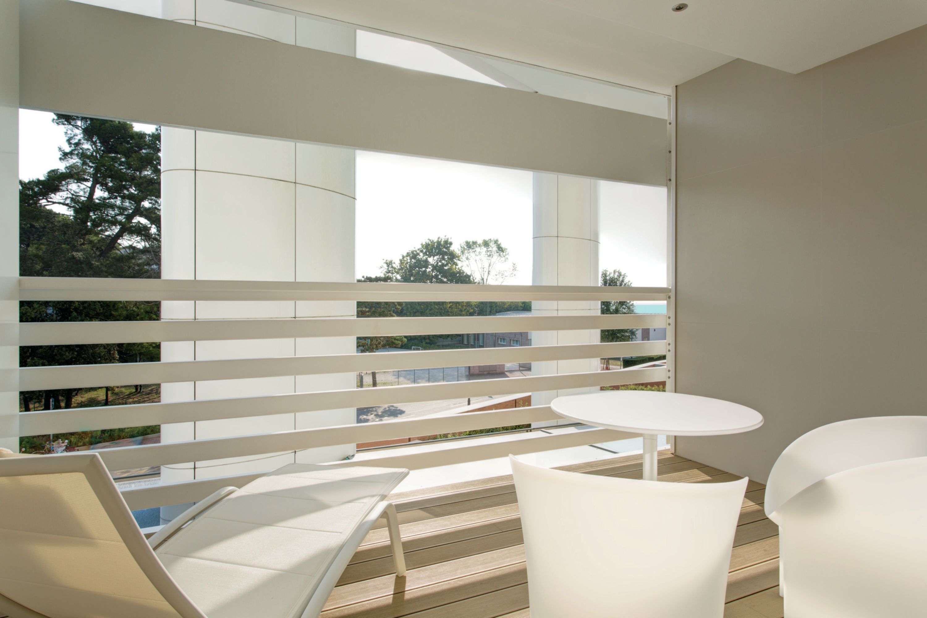 A modern balcony with white furniture, large windows, and a view of the cityscape.