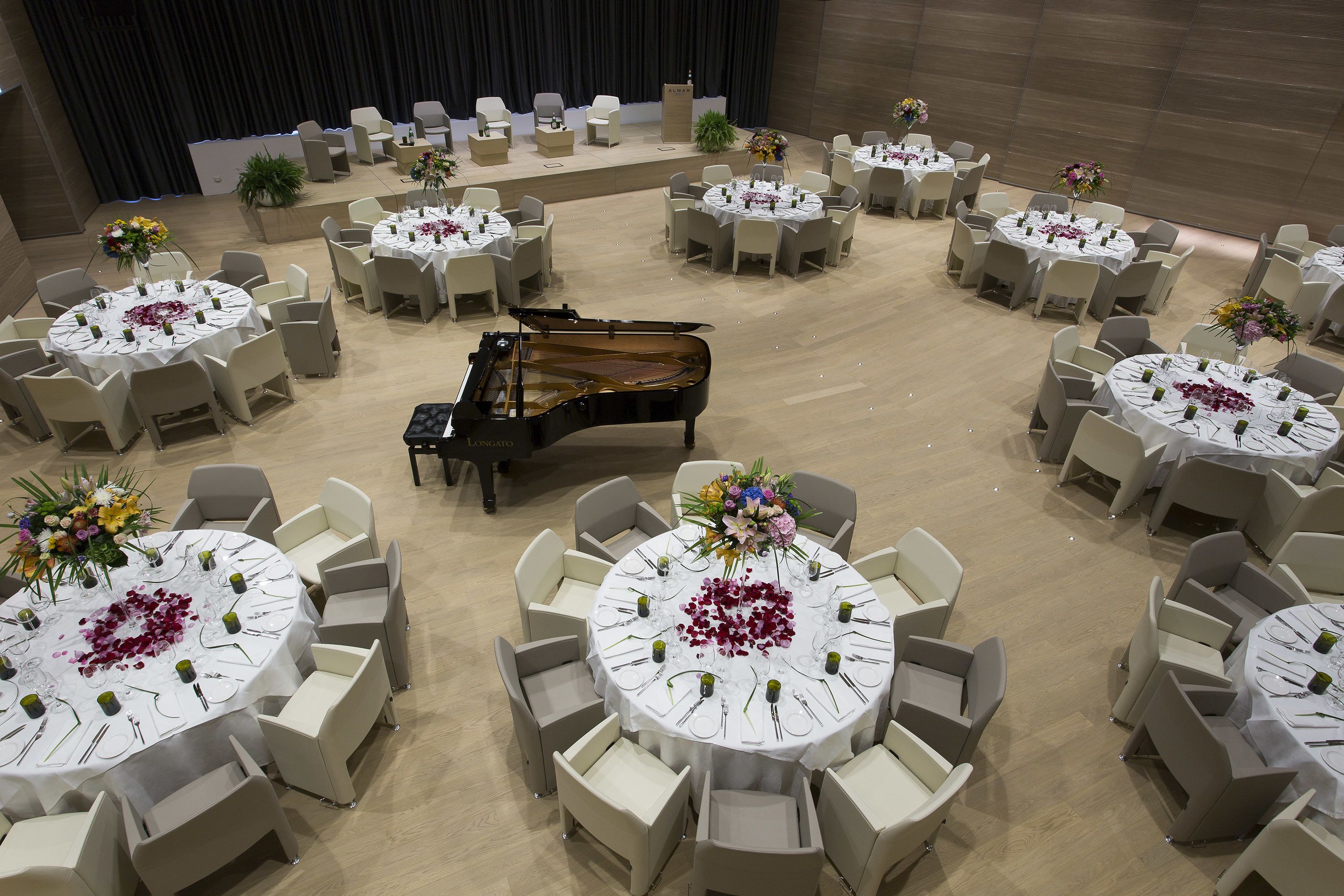 Un salón de banquetes con mesas redondas, cada una con mantel blanco, sillas y centros de mesa florales. Un piano de cola se encuentra ubicado en el centro.