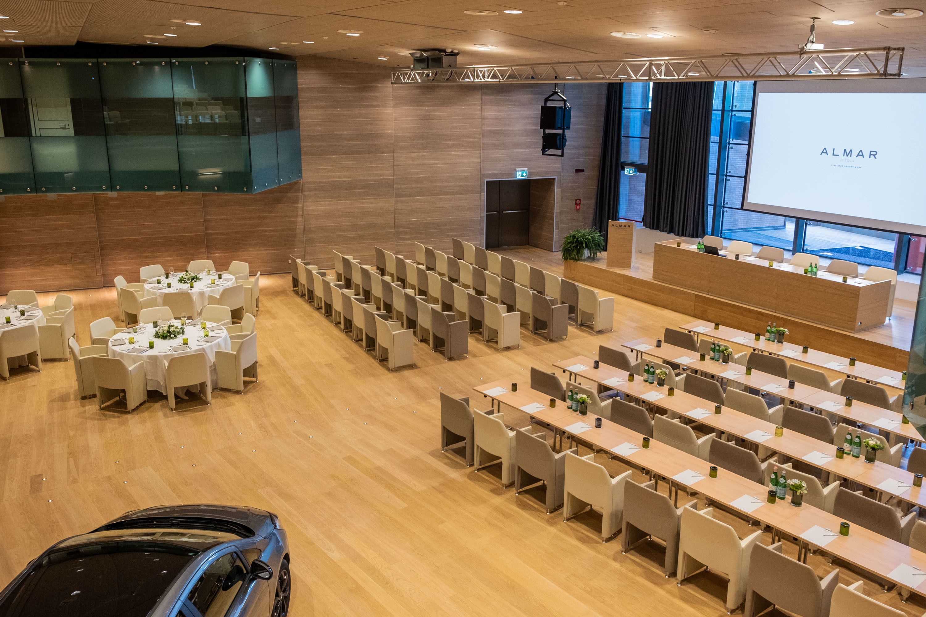 A conference room with rows of tables and chairs, a stage, and a large screen displaying the word "ALMAR".