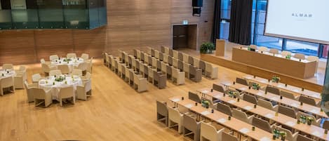 A conference room with rows of tables and chairs, a stage, and a large screen displaying the word "ALMAR".