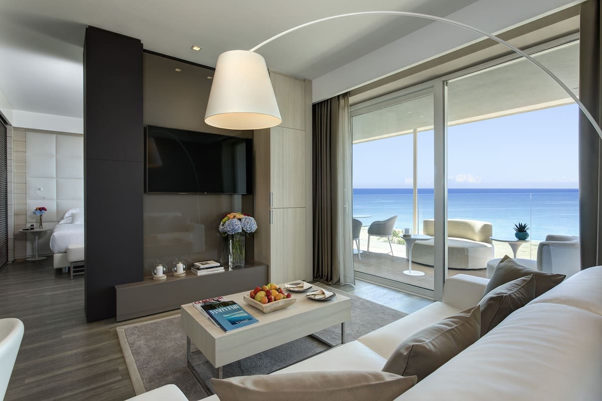 A modern hotel room with a large window offering a sea view, a flat-screen TV mounted on the wall, and a comfortable seating area with a sofa and armchairs.