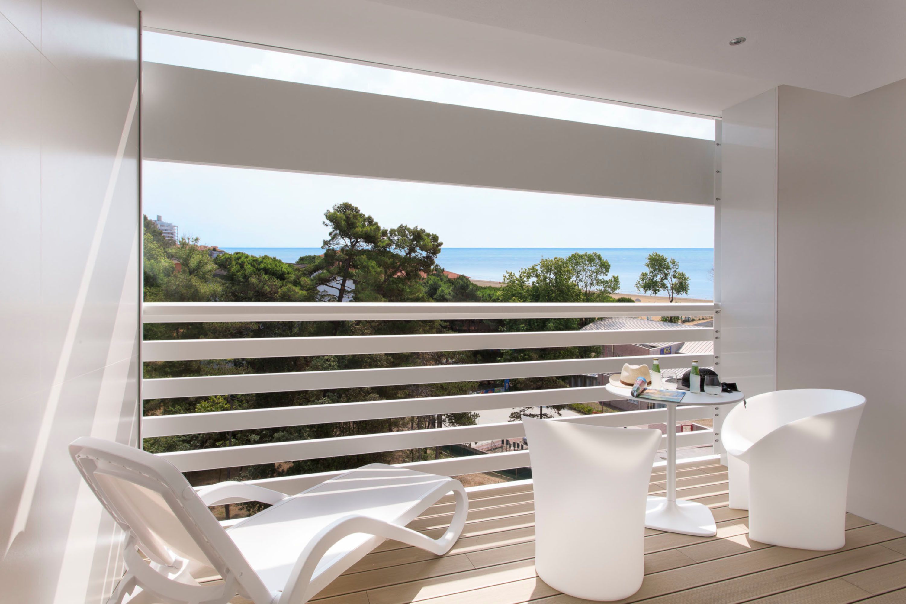 A balcony with white chairs and a table, overlooking a beach with trees and buildings.