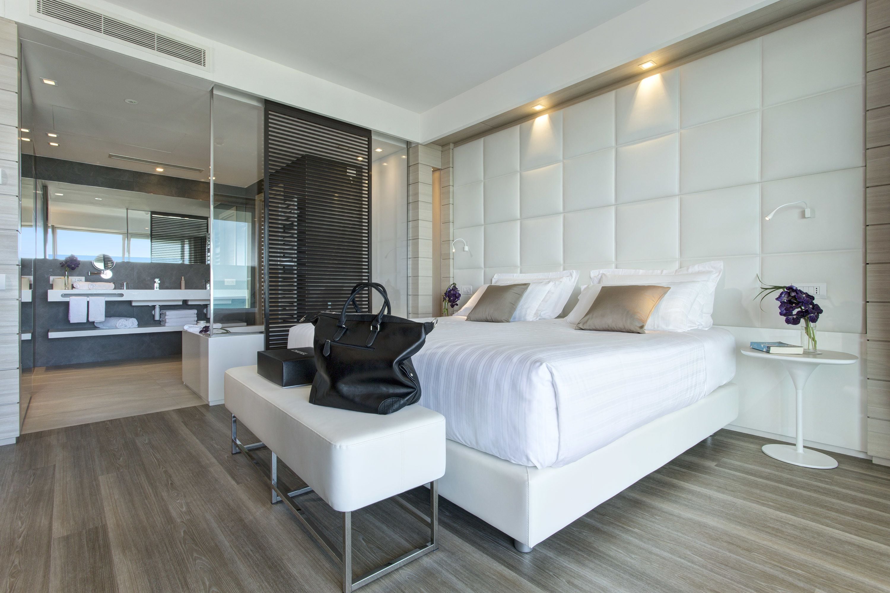 A modern bedroom with a large bed, a bench, a bedside table, and a mirror.