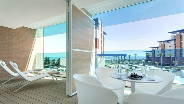 A modern balcony with white furniture, a glass partition, and a view of the ocean and buildings. - Almar Lido Jesolo Resort & Spa (Jesolo)