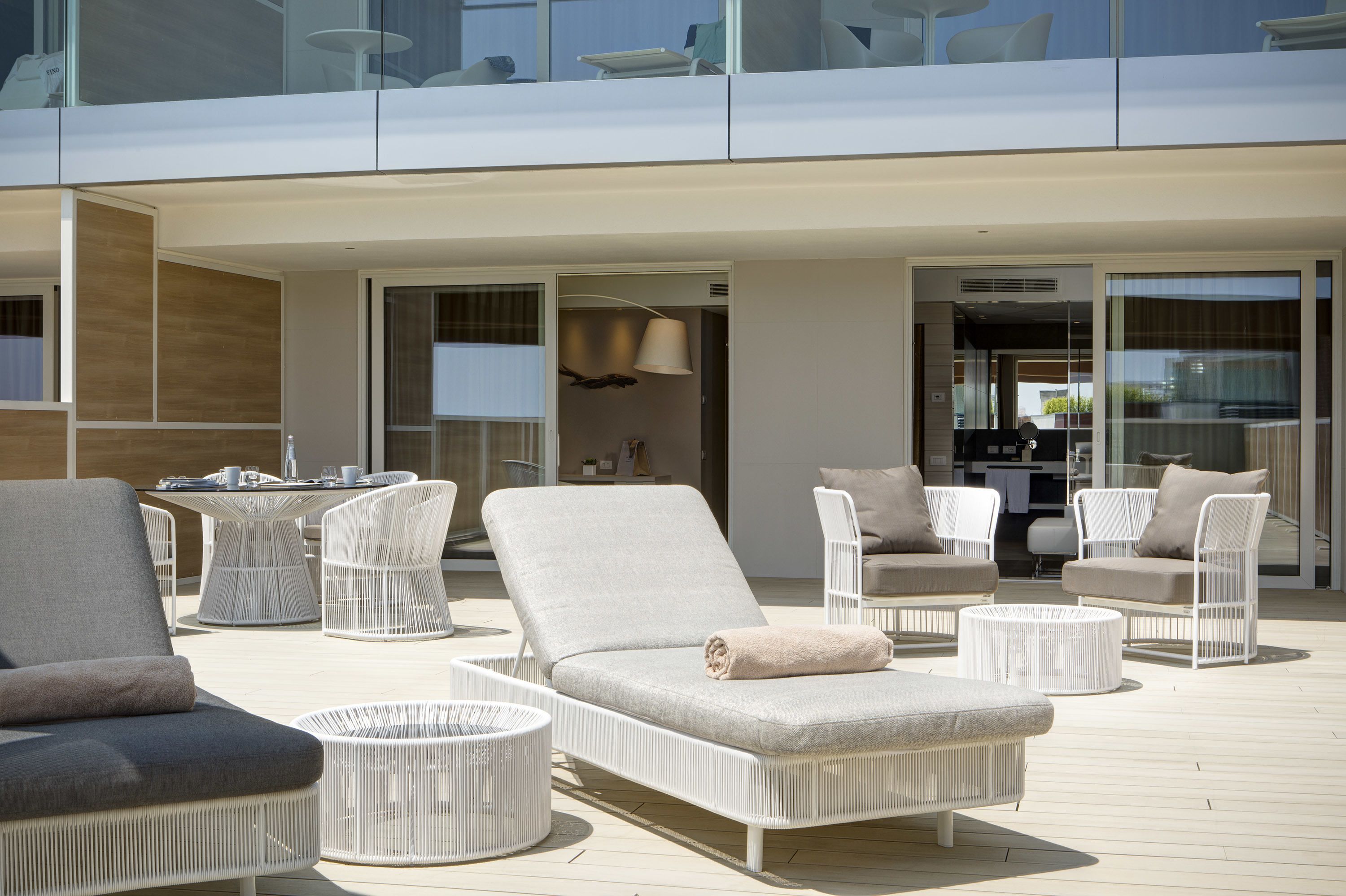 Ein moderner Außenbereich mit Lounge-Möbeln aus weißem Rattan, darunter Liegestühle und ein runder Tisch, sowie ein hintergrundseitiger Küchenbereich mit Glasfassade.