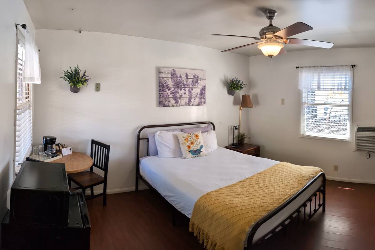 A bedroom with a bed, a desk, a chair, a ceiling fan, a window with blinds, and a wall-mounted air conditioner.