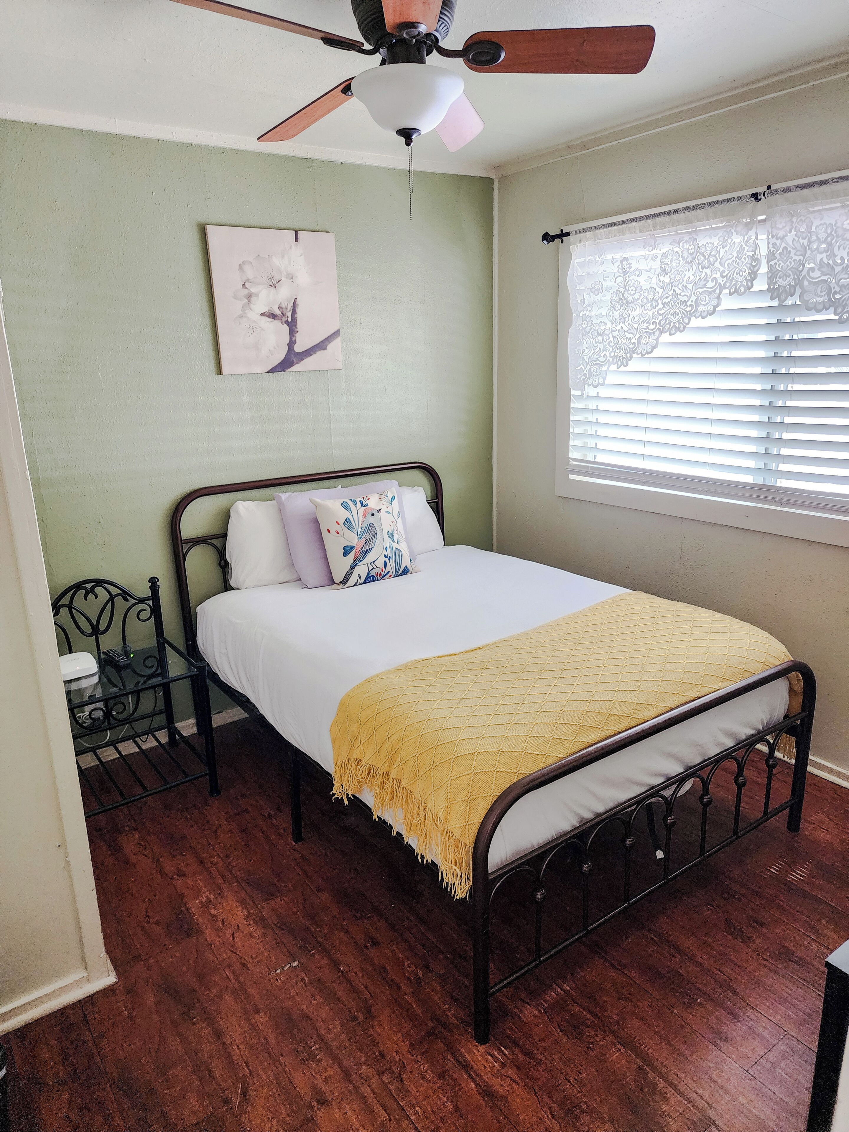 Une chambre à coucher avec un lit, un ventilateur de plafond, une fenêtre avec des stores et un tableau accroché au mur.