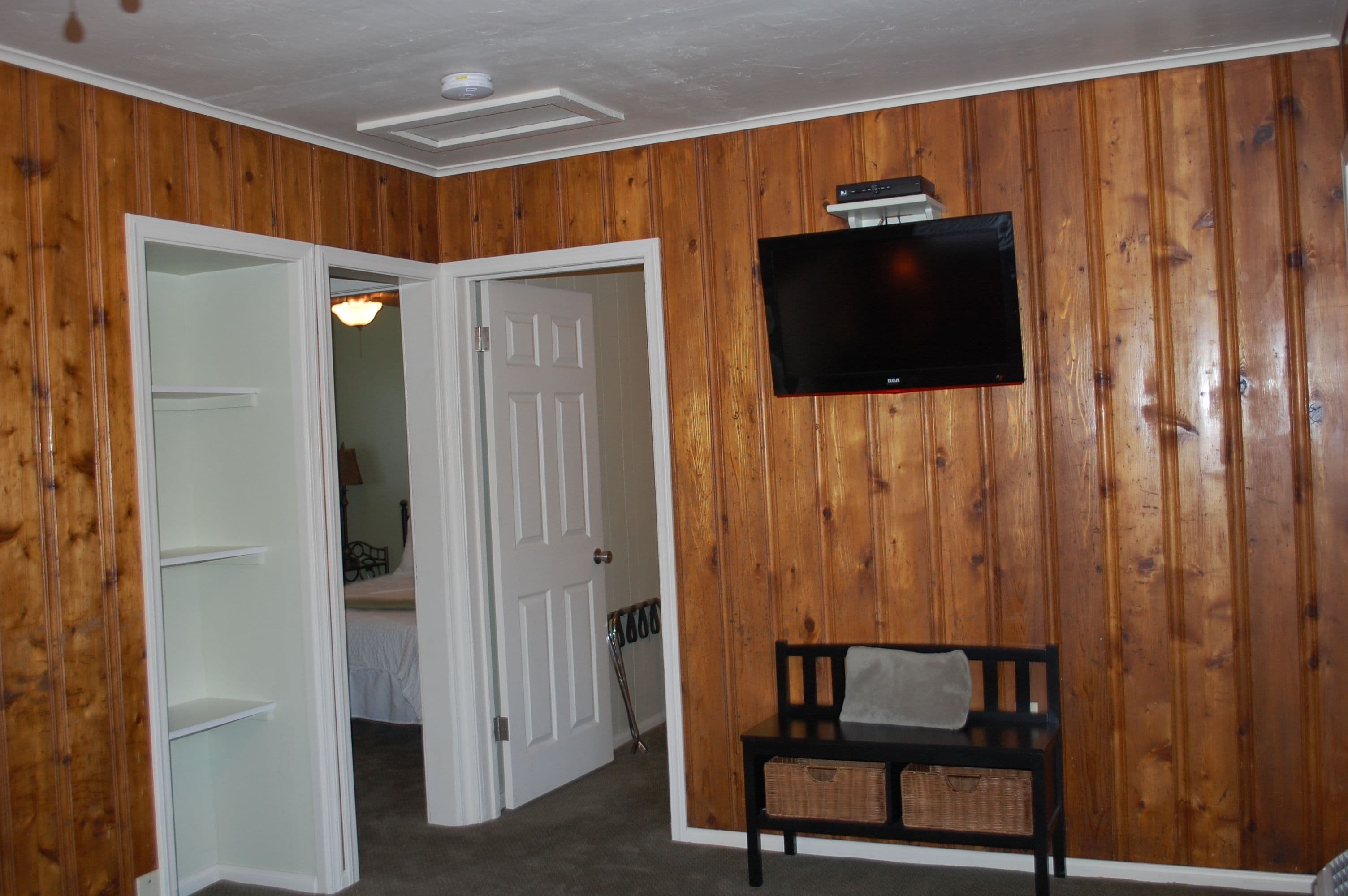 A room with wooden panel walls, a flat-screen TV mounted above a bench, and a door leading to another room.
