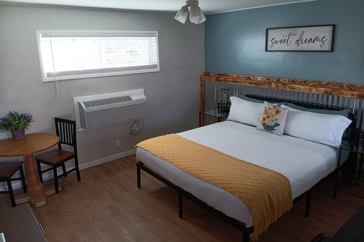 A bedroom with a bed, a window, a ceiling fan, a wall-mounted air conditioner, a small table with a chair, and a potted plant.