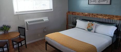 Un dormitorio con cama, ventana, ventilador de techo, aire acondicionado de pared, una mesita con silla y planta en maceta.